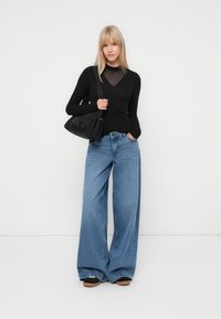 Black long-sleeve top with a mesh neckline, paired with light blue wide-leg jeans and a black fabric bag. Textured fabric and relaxed fit.