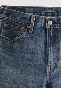 Calças de ganga em lavagem escura com ferragens em cobre, apresentando um design de cinco bolsos, costuras visíveis e fecho com botão metálico.