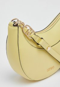 Sac à main en cuir jaune de forme incurvée, avec ferrures dorées et coutures noires contrastantes. Comprend une fermeture éclair et une sangle réglable.