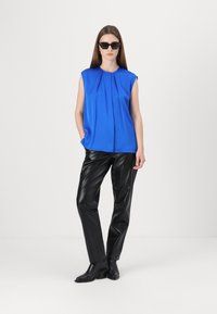 Blusa senza maniche blu con pieghe frontali, abbinata a pantaloni in ecopelle nera e stivali alla caviglia. La modella indossa occhiali da sole neri.