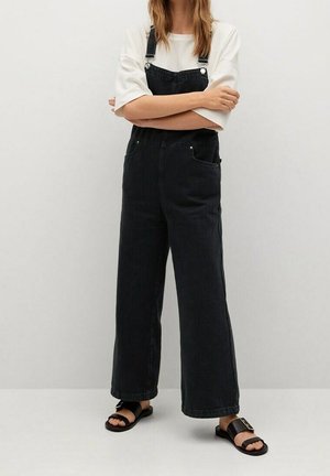 Nainen, joka seisoo käsivarret ristissä valkoinen t-paita, mustat leveälahkeiset farkkukokonaisasut ja mustat avoimet sandaalit päällään yksiväristä seinää vasten.