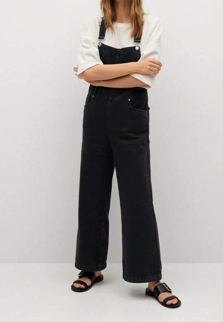Femme aux bras croisés portant un t-shirt blanc, une salopette large en denim noir et des sandales noires à bout ouvert, debout devant un mur uni.