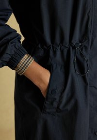 Geacă de vânt de marină cu manșete elastice, talie cu șnur și buzunare laterale. Accesoriu din material în carouri pe manșeta interioară. Material neted și ușor.