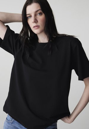 Jeune femme aux longs cheveux noirs portant un t-shirt noir ample et un jean bleu, posant avec un bras derrière la tête.