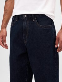 Bărbat îmbrăcat în blugi de denim bleu închis și un tricou alb, arătându-și mâinile și tatuajul de pe antebrațul stâng pe un fundal simplu.