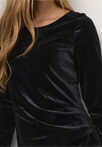 Černý sametový top s dlouhými rukávy a zaobleným výstřihem, s drapériovým designem a hladkou texturou.