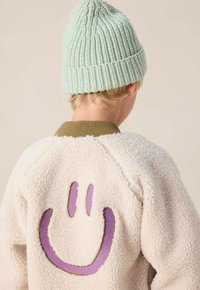 Kind in een crèmekleurige fleecejack met een paarse glimlach op de achterkant en een lichtgroene gebreide muts, gezien van achteren.