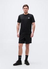 Νέος άνδρας που στέκεται, φορώντας μαύρα αθλητικά ρούχα Adidas: T-shirt, σορτς, κάλτσες και αθλητικά παπούτσια, με φόντο απλό.