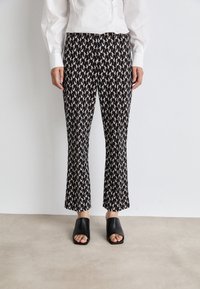 Black patterned flared trousers with a repeating abstract design in beige and cream tones, paired with chunky black open-toe slides.