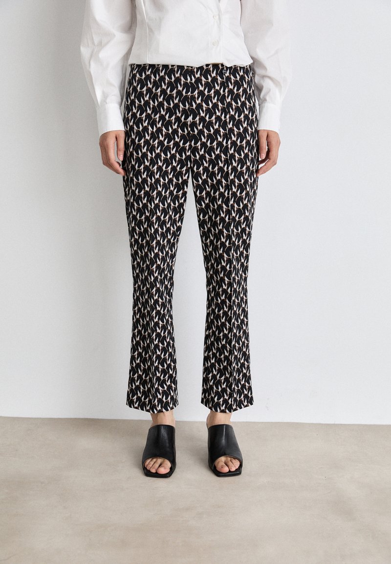 Black patterned flared trousers with a repeating abstract design in beige and cream tones, paired with chunky black open-toe slides.
