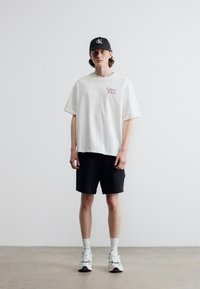 Νεαρός άνδρας που φοράει λευκό μπλουζάκι Calvin Klein, μαύρο σορτς, λευκές κάλτσες, αθλητικά παπούτσια και μαύρο καπέλο Calvin Klein, στέκεται μπροστά σε απλό φόντο.