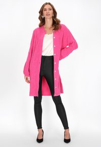 Cardigan lavorato a maglia di un vivace rosa acceso con un motivo a trecce, dotato di bottoni argentati e una vestibilità comoda, abbinato a pantaloni neri attillati e tacchi.