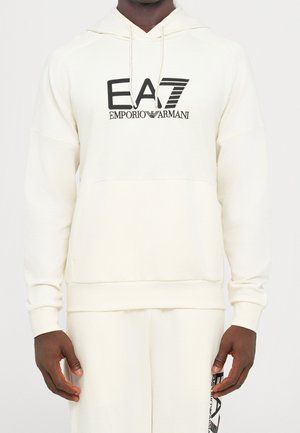 Mężczyzna ubrany w jasnobeżową bluzę z kapturem EA7 Emporio Armani i dopasowane spodnie z czarnym logo na klatce piersiowej i udzie, stojący z rozluźnionymi ramionami.