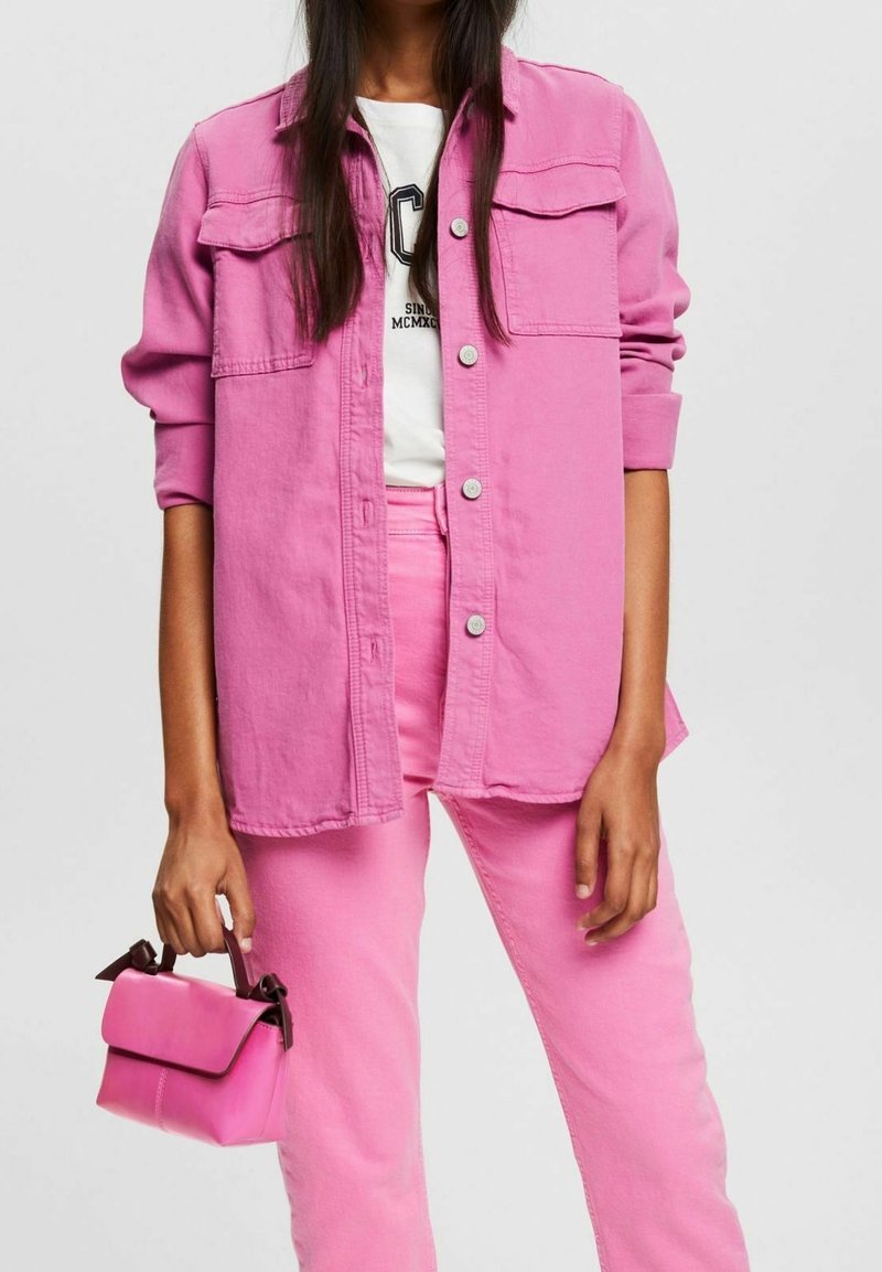 Różowa dżinsowa kurtka z guzikami i dwiema kieszeniami na piersi, zestawiona z pasującymi różowymi dżinsami oraz małą różową torebką ozdobioną wstążkami.