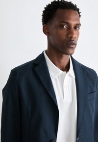 Sacou texturat bleu marin cu un singur nasture, purtat peste un tricou polo alb cu guler clasic și clapa cu trei nasturi.