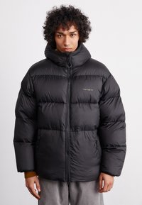 Schwarze Daunenjacke mit hohem Kragen, Reißverschluss und elastischen Bündchen. Verfügt über eine strukturierte Oberfläche und dezente Markenkennzeichnung auf der Brust.