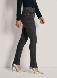 Persoon draagt aansluitende zwarte jeans en zilveren hoge hakken, staat zijwaarts tegen een lichte achtergrond.