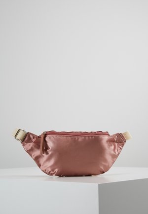 Sac banane mauve à texture satinée avec une fermeture éclair supérieure et une sangle ajustable beige, posé sur un socle blanc sur fond gris.