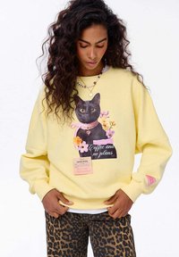 Žena s dlouhými kudrnatými vlasy, oblečená v žluté mikině s černou kočkou, květinami a textem "Káva a žádné plány", v kombinaci s kalhotami s leopardím vzorem.