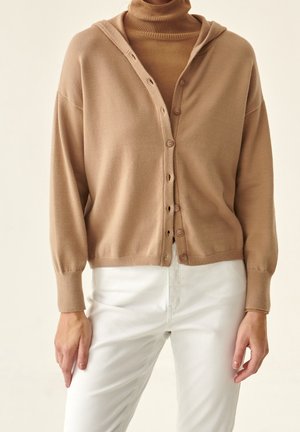 Kobieta w beżowym swetrze na guziki noszonym na beżowym golfie, w białych spodniach, stojąca na tle o neutralnym kolorze.