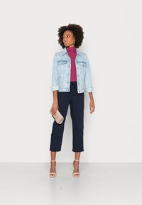 Veste en denim bleu clair, col roulé violet, pantalon cropped bleu marine, talons nus et un clutch rose. Comprend des manches retournées et des détails de poche.