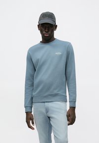 Ανοιχτό μπλε φούτερ με στρογγυλή λαιμόκοψη, ριγκάκια στα μανίκια και μικρό λευκό λογότυπο στο στήθος, συνδυασμένο με ανοιχτόχρωμο τζιν και σκουρόχρωμο καπέλο.