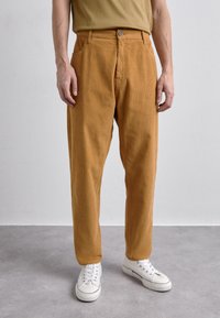 Pantalones de pana en color beige con un corte relajado, que cuentan con un cierre frontal de botón y cremallera, bolsillos laterales y zapatillas blancas.