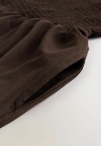 Brunt stof med en blød tekstur har et rynket design og opsamlet detalje ved sømmen, hvilket skaber en flydende form.