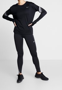 Persoon in een zwart Nike lange mouwen top en legging met grijze accenten, staand met handen op de heupen en zwarte sport schoenen.