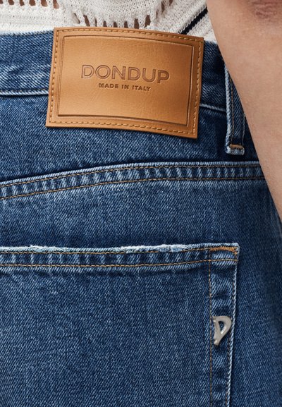 Jean en denim bleu avec un patch en cuir marron portant l'inscription "DONDUP MADE IN ITALY" au-dessus de la poche arrière et une petite étiquette métallique en forme de "D".
