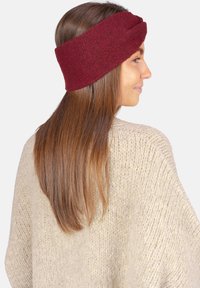 Bandeau tricoté bordeaux avec un design torsadé, en matière douce, porté sur de longs cheveux, associé à un pull épais de teinte claire.