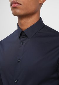 Camisa de botones azul marino con cuello estructurado, botones brillantes y textura de tela suave, que presenta un corte entallado y un diseño minimalista.