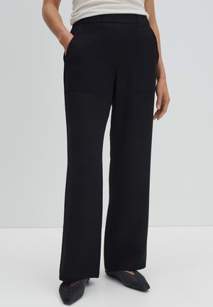 Vrouw die zwarte broek met brede pijpen en voorzakken draagt, samen met zwarte puntige platte schoenen, staand met één hand in de zak.