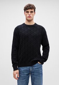 Maglione nero lavorato a maglia con un motivo testurizzato, scollo rotondo e bordi a costine. Abbinato a jeans in denim azzurro chiaro.