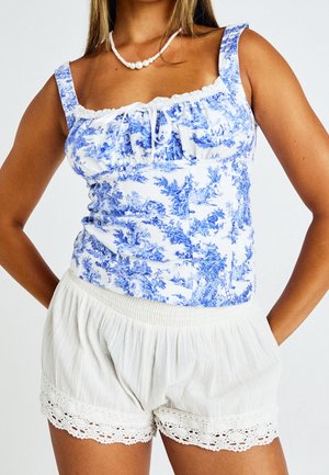 Femme portant un haut sans manches à motifs bleus et blancs avec un nœud blanc, un short élastique blanc avec un bord en dentelle et un collier de perles.