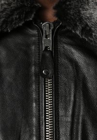 Černá kožená bunda se šedým kožešinovým límcem, s stříbrným zipem a koženým jezdcem. Detailní záběr na zip.