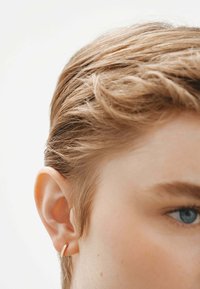 Pendiente de aro dorado exhibido en la oreja izquierda, iluminando la textura suave del metal contra el cabello rubio claro y texturizado y la piel clara.