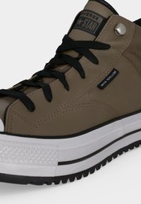 Καφέ ψηλά sneakers Converse με μαύρα κορδόνια, λευκή υφασμάτινη σόλα, μεταλλική τρύπα και ετικέτα αδιαβροχοποίησης στη πλευρά.