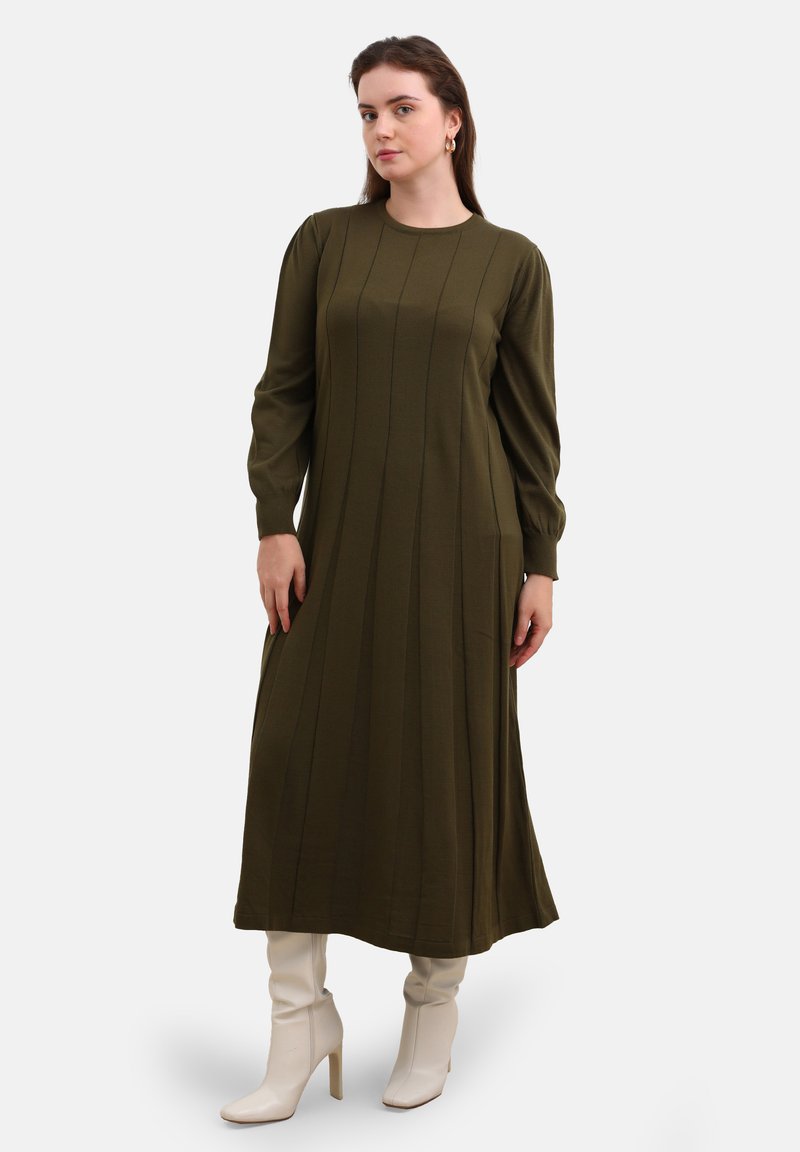 Olivgrünes langärmliges Kleid mit vertikalen Nähten, rundem Halsausschnitt und schwingendem Saum, kombiniert mit weißen Stiefeletten mit Absatz. Weiche Stofftextur.
