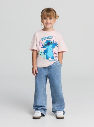 Maglietta di cotone rosa con grafica di un personaggio dei cartoni animati blu, abbinata a jeans larghi in denim azzurro chiaro e sneakers bianche.