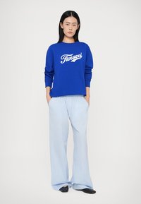 Hanorac albastru cu logo-ul "Fiorucci" alb, asortat cu pantaloni largi, cu dungi albastre deschise. Modelul stă în picioare cu mâinile în buzunare, purtând pantofi negri.