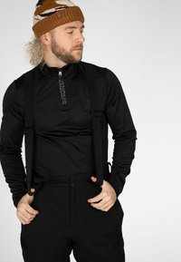 Top funzionale nero con mezza cerniera e logo, abbinato a pantaloni neri con straps regolabili. Cappello lavorato a maglia con motivo marrone e bianco.