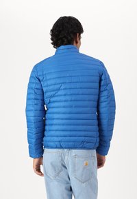 Blauwe gevoerde jas met horizontale stiksels, hoge kraag, elastische manchetten en een gladde textuur. Draag over lichtblauwe spijkerbroeken.