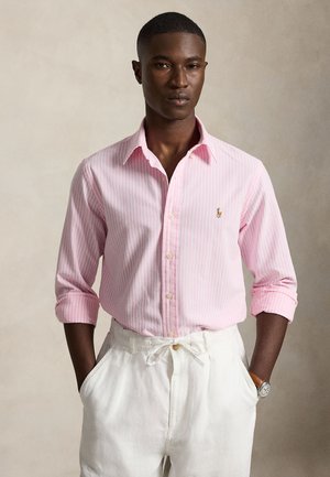 Jeune homme portant une chemise à rayures rose clair à manches retroussées et un pantalon blanc à cordon, debout devant un fond uni.