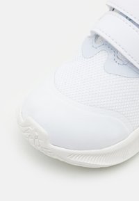 Zapatilla deportiva blanca con parte superior de malla, puntera de goma suave y correas de velcro. Suela texturada con un patrón de estrellas en el talón.