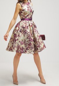 Nainen pukeutuneena hihattomaan, polvipituiseen kukkakuviolliseen mekkoon, jossa on violetteja sävyjä, beiget korkokengät ja hän kantaa tummanviolettia clutch-laukkua.