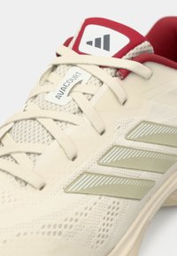 Beżowy sportowy but Adidas Avacourt z kremowymi sznurowadłami, oliwkowymi paskami po bokach i czerwonym, wyściełanym kołnierzem.