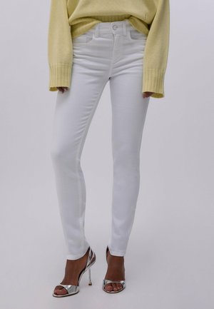 Mujer con jeans blancos ajustados, suéter amarillo de manga larga y sandalias plateadas de tacón alto, de pie frente a un fondo liso.