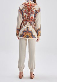 Veste beige à motifs avec un design paisley coloré, cintrée à la taille. Associée à un jogging crème, avec des poignets élastiques. Sandales à talons en bois.