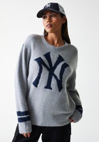 Grijze gebreide trui met een marineblauw NY-logo, geribbelde halslijn en gestreepte mouwen. Zachte textuur, relaxte pasvorm en verlaagde schouders voor comfort.